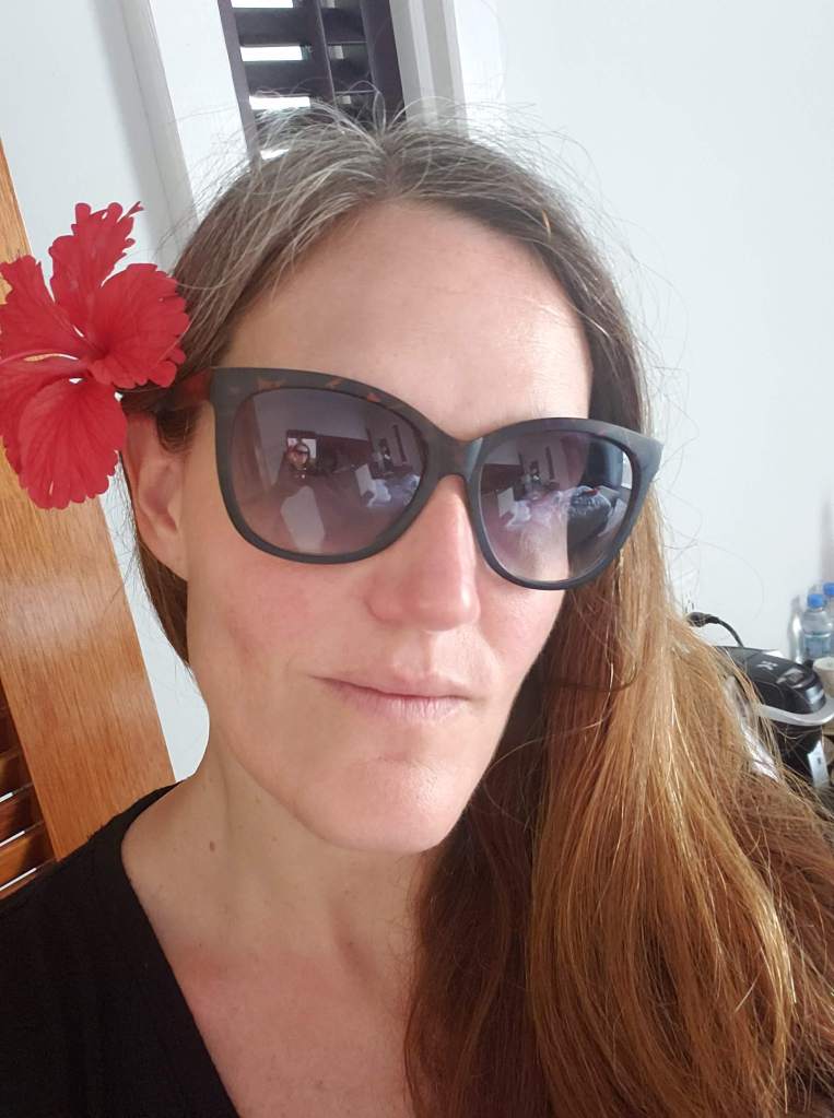 Photo of a white woman with gray-brown hair and a red flower tucked behind her ear. She is wearing a black shirt and dark sunglasses.